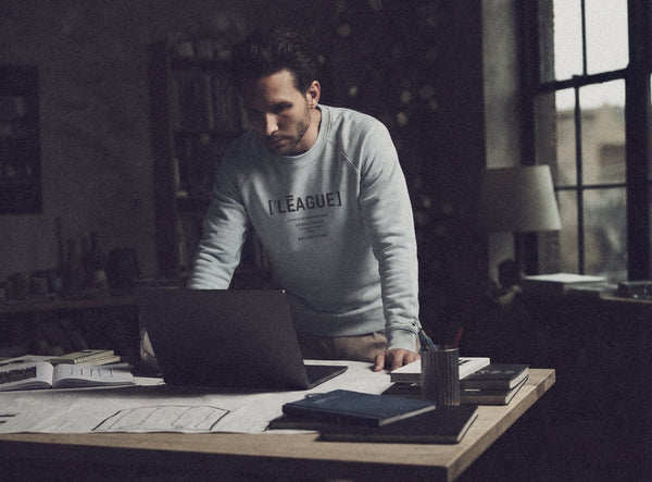 Man wearing a blue ['LĒAGUE] sweatshirt in an creative office setting. 