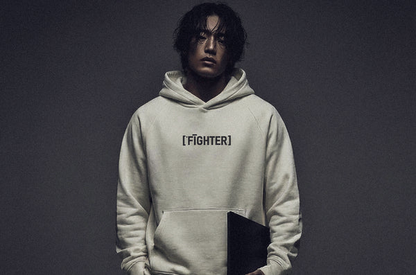 Model wearing ['FĪGHTER]™ hoodie in dark tone, minimal studio background