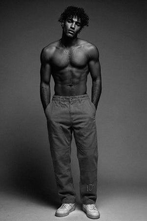 Man standing shirtless wearing dark pants against a gray background