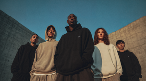 Models wearing oversized hoodies from the ['INITIĀTION] COLLECTION by ['LĒAGUE] standing together in a modern architectural setting