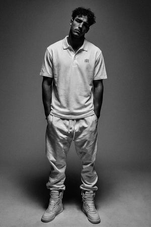 Man wearing a light-colored polo shirt and pants with a logo, standing against a gray background