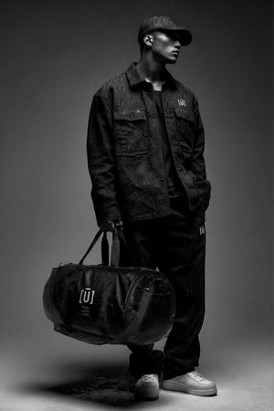 Person wearing a dark outfit with a cap, holding a duffel bag on a gray background