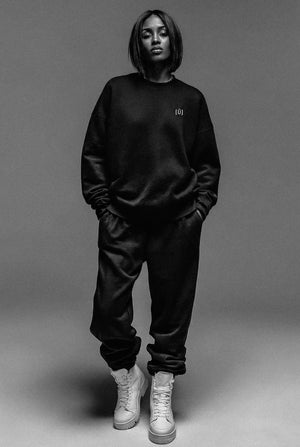 Person wearing a black sweatshirt and pants with a logo, standing against a gray background