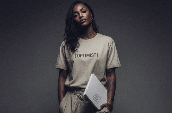 Model wearing ['OPTIMIST]™ t-shirt in soft neutral tone, minimal studio setting